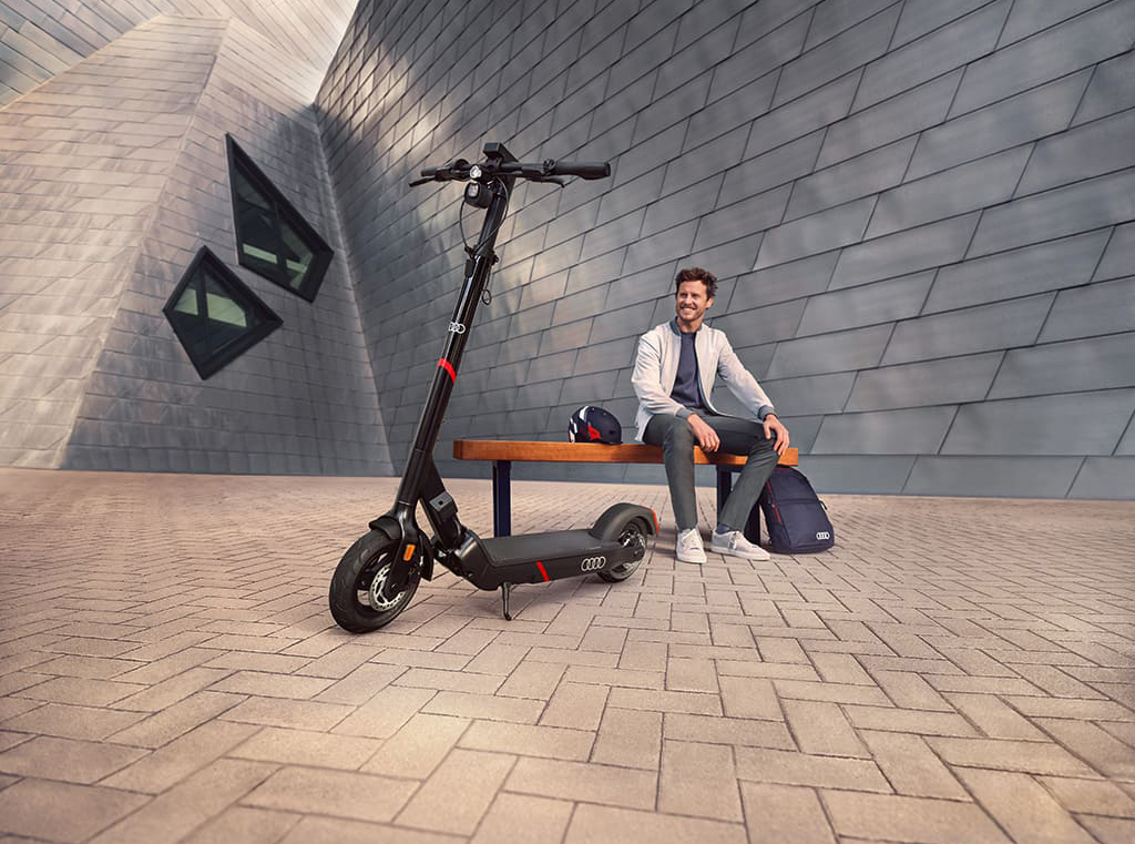 Audi Original E-Scooter im Vordergrund; Mann sitzend auf einer Bank vor einem Gebäude im Hintergrund